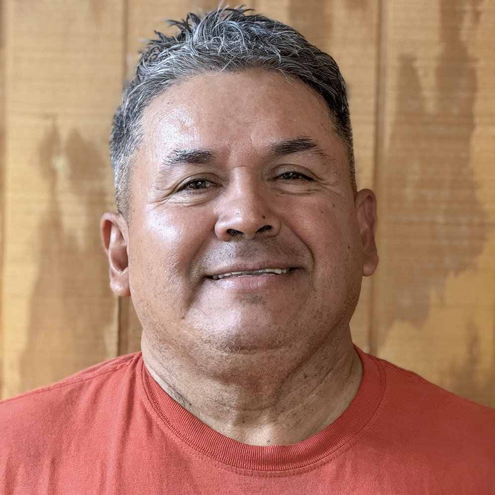 Carlos San Juan of Coastal Fumigators is shown standing in front of a wood paneled wall smiling. He has salt and pepper hair and is wearing a red shirt.