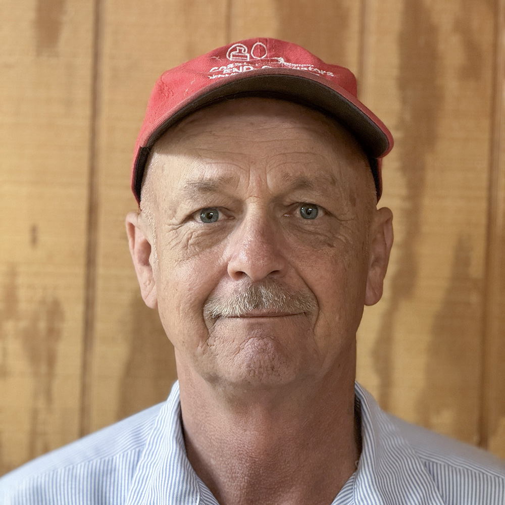 Denny Clifton of Coastal Fumigators is standing in front of a wood paneled wall smiling. He is wearing a blue and white striped button up shirt and a red baseball cap.