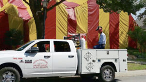 A man is shown sitting in the bed of a white Coastal Fumigators truck monitoring the fumigation process. There is a home tented in a yellow and red striped fumigation tent in the background.