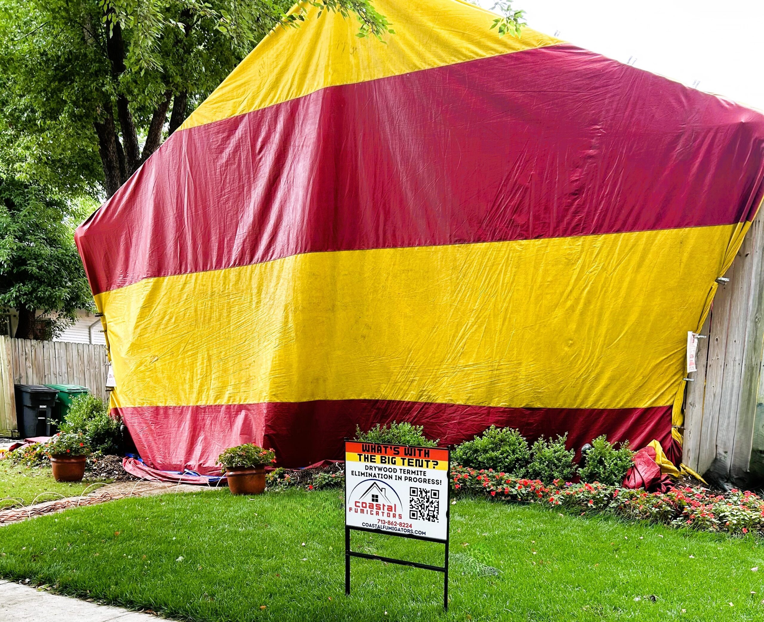 A red and yellow fumigation tent is over a home surrounded by green grass and flowers and a commercial sign.