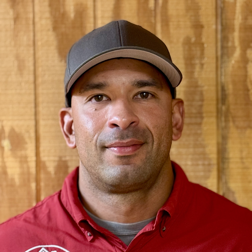 Carlos San Juan of Coastal Fumigators is shown standing in front of a wood paneled wall smiling. He has salt and pepper hair and is wearing a red shirt.