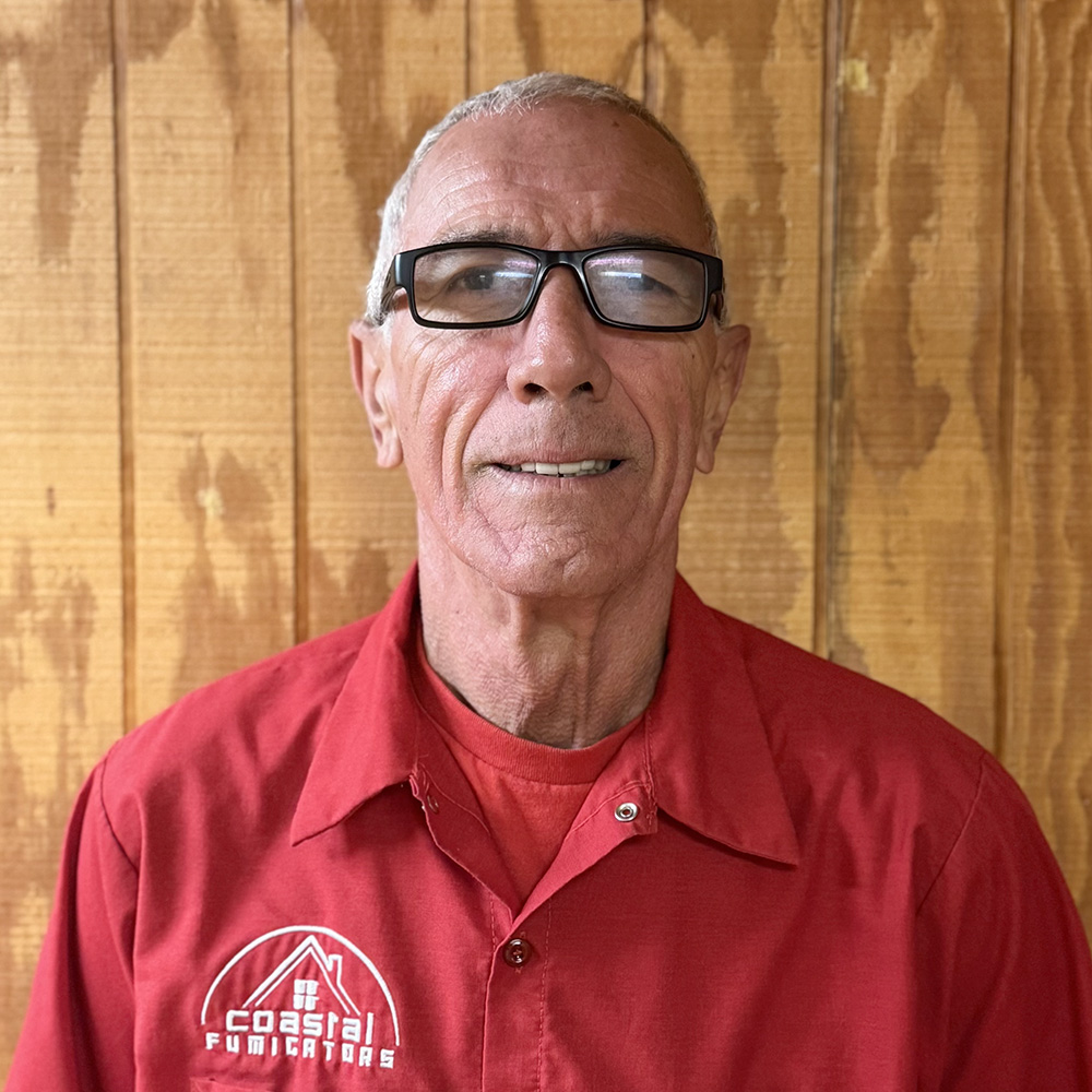 Denny Clifton of Coastal Fumigators is standing in front of a wood paneled wall smiling. He is wearing a blue and white striped button up shirt and a red baseball cap.